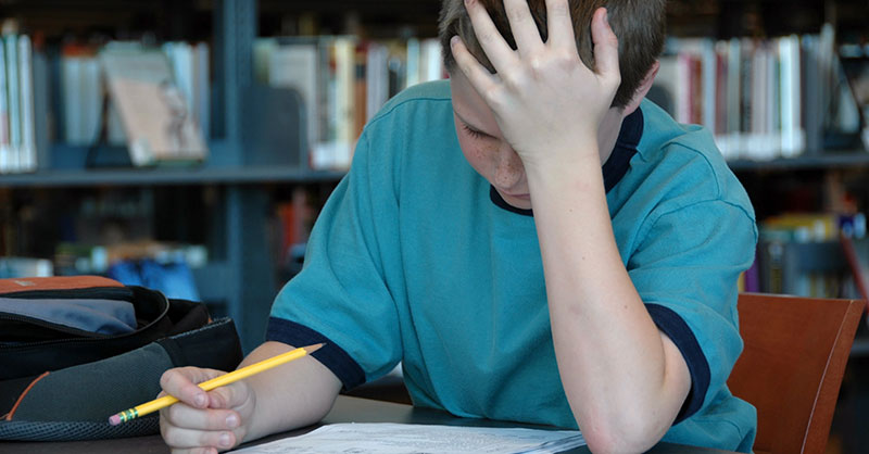 A teenage boy sits at a table in front of a bookshelf. He is hunched over a piece of paper. He holds a pencil in his right hand, and his left hand presses against his forehead.