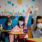 Children in classroom seated at their desks, raising their hands.