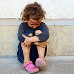 A young girl sits against a building, holding her knees and looking down.