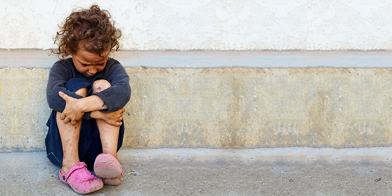 A young girl sits against a building, holding her knees and looking down.