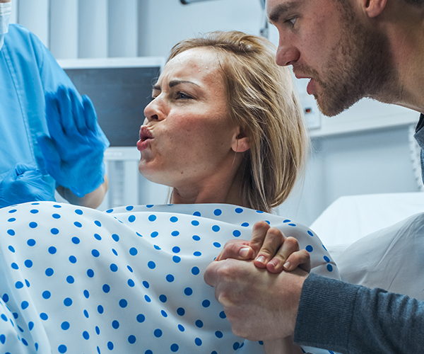 A blonde pregnant woman in labor breathes heavily, while her husband holds her hand and a masked healthcare worker looks on.