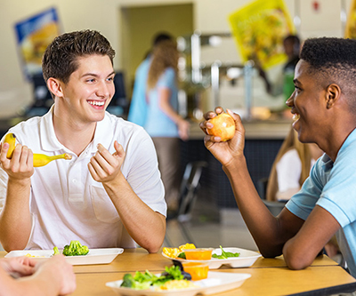 Two smiling teenage boys, one holding a banana and one holding an apple, sit at a table filled with plates of vegetables.