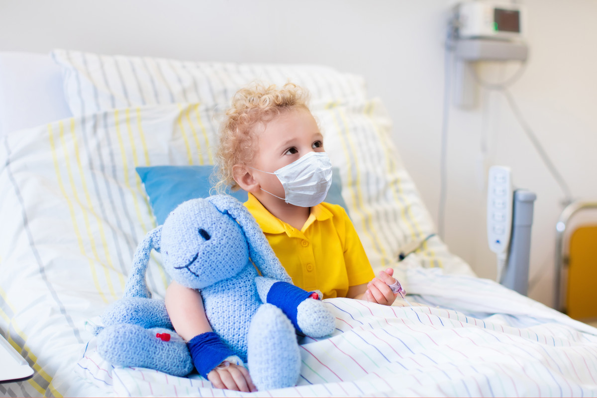 A boy sits up in a hospital bed. He’s wearing a mask and holding onto a blue stuffed bunny.