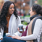 A Black woman and her pregnant White friend sit on a park bench having coffee and talking.