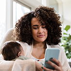 A mother holding an infant while also looking at a smartphone.