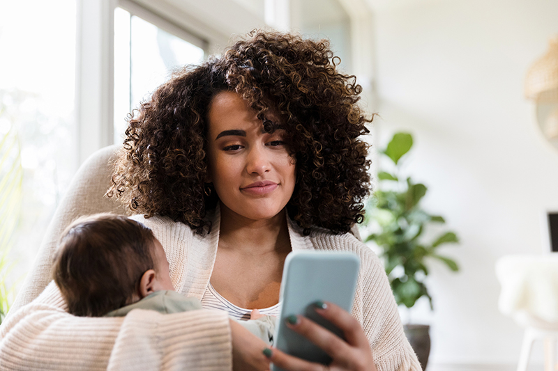 A mother holding an infant while also looking at a smartphone.