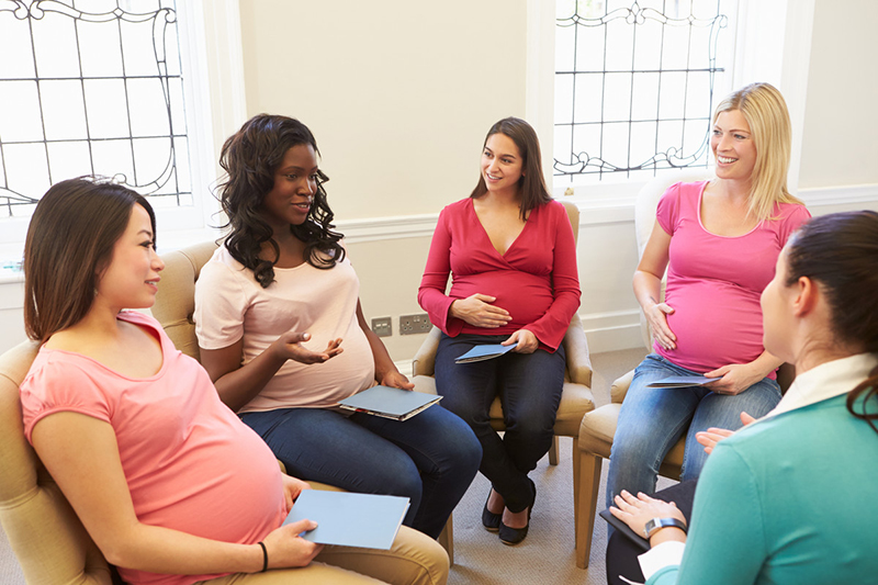 Five pregnant women sit in a circle talking with two large windows in the background.
