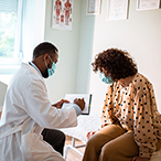 Patient seated on examination table facing medical professional holding a clipboard.