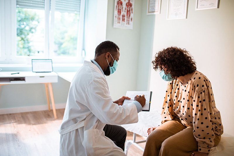 Patient seated on examination table facing medical professional holding a clipboard.