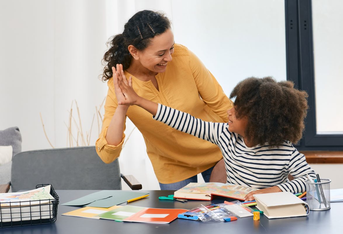 Adult and preteen high five behind desk with books folders, pens and pencils.