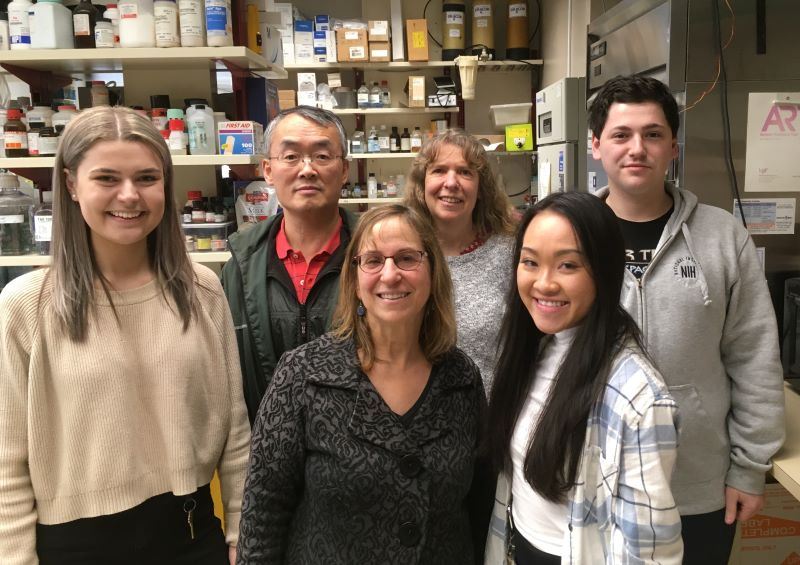 Six people stand in a laboratory in front of shelves that contain various bottles and boxes.