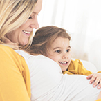 A pregnant woman wearing a white shirt and yellow cardigan lies on a couch with her toddler daughter, whose hand is placed on her mother’s belly.