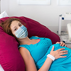 Pregnant person, masked, on their back in a hospital setting.