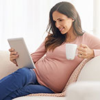 A pregnant woman sits on a sofa reading a tablet device and holding a mug in her hand.