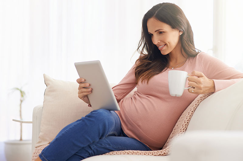 A pregnant woman sits on a sofa reading a tablet device and holding a mug in her hand.