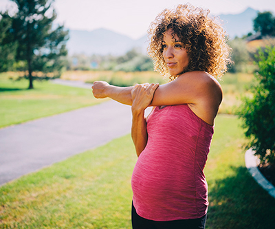 A pregnant woman in a pink shirt stretches her shoulder in a park with a jogging path.