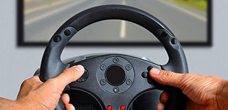 Hands on steering wheel in front of a computer monitor.