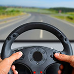 Hands on steering wheel in front of a computer monitor.