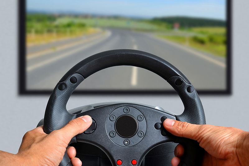 Hands on steering wheel in front of a computer monitor.