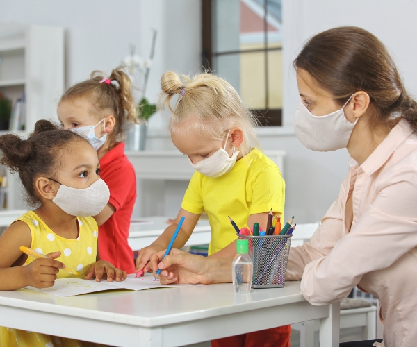 Young children are seated at a table and drawing with a teacher. Everyone is wearing facemasks.