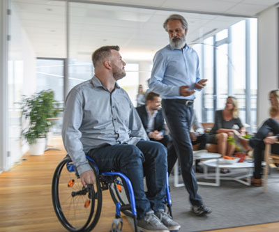 A man in a wheelchair has a conversation with another man who is standing. Both are moving across a room where other adults are seated around a coffee table.