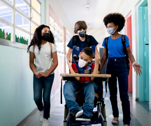 A group of teenagers walk down a school hallway, and one student is being pushed down the hall in his wheelchair. Everyone is wearing facemasks.