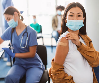 A pregnant woman shows off her post-vaccination band-aid for the camera. A healthcare provider and other patients are visible in the background. Everyone is wearing facemasks.