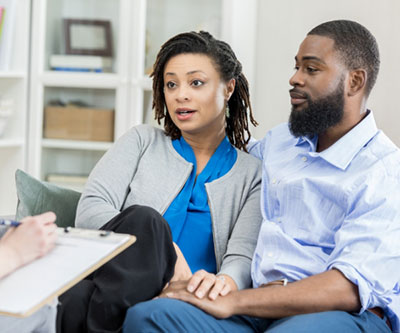 A couple is seated and holding hands in front of a partially visible healthcare provider who is writing on a clipboard.