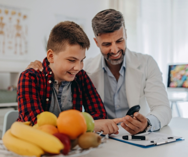 A healthcare provider checks a teenager’s blood glucose by using a fingerprick on a monitor. Both are smiling, and a plate of fresh fruit is visible on the table.