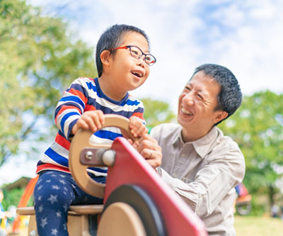 A father and son, who has Down Syndrome and is wearing glasses, are playing together in an outdoor playground. Both are smiling