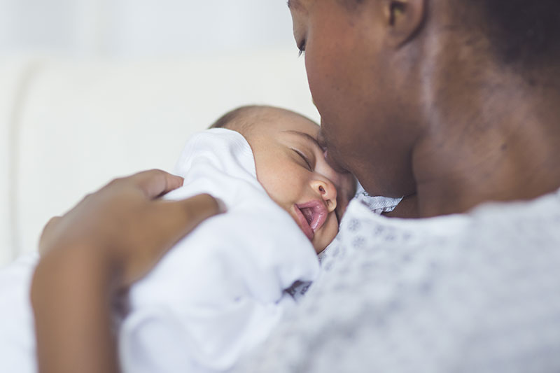 A Black woman wearing a hospital gown kisses the forehead of her newborn baby.