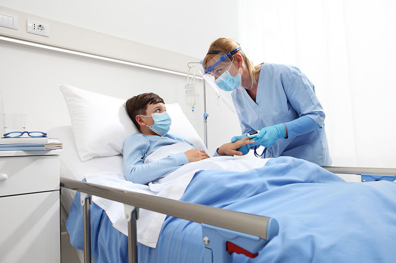 Masked child in hospital bed with masked medical professional at bedside.