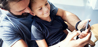 A man and a smiling little boy sitting in his lap look at a mobile phone.