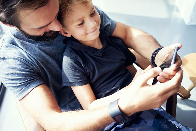 A man and a smiling little boy sitting in his lap look at a mobile phone.