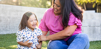A little girl with Down syndrome sits on the grass with her adult female caretaker. Both are laughing. Trees and a stone wall are visible in the background.