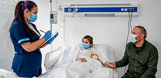 Child in hospital bed, health care worker at left writing on clipboard, parent seated at right. All are masked.