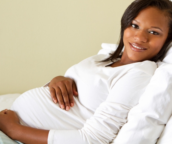A smiling pregnant woman holds her belly while reclined in bed.