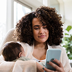 A mother holding an infant and looking at a smartphone.