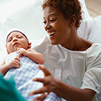 A smiling Black woman wearing a hospital gown opens her arms to hold a newborn wrapped in a blue and white blanket. A healthcare worker wearing green scrubs is handing her the baby.