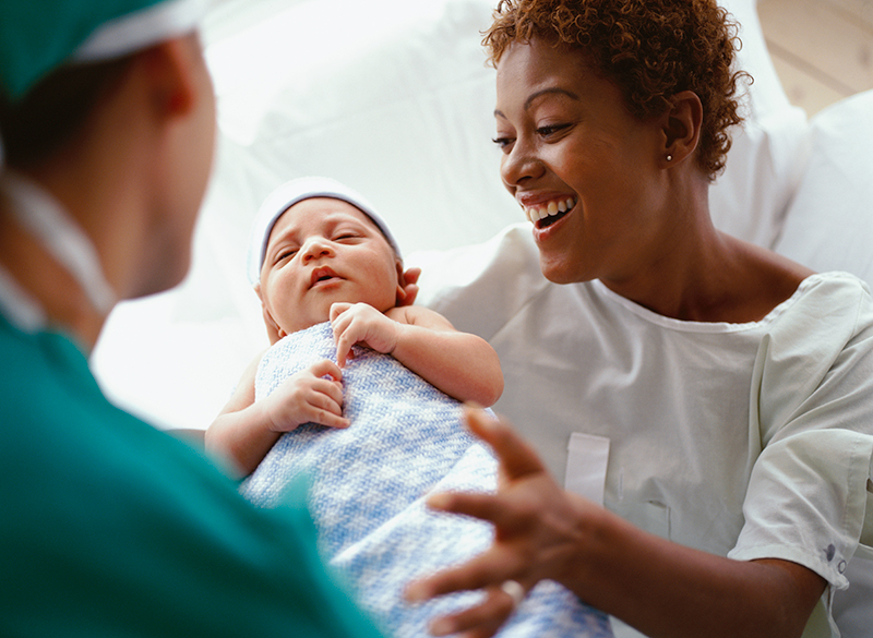 A smiling Black woman wearing a hospital gown opens her arms to hold a newborn wrapped in a blue and white blanket. A health care worker wearing green scrubs is handing her the baby.