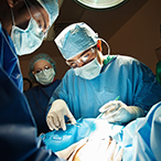 Medical professionals in surgical garb surround the draped torso of a pregnant person, while another medical professional holds surgical instruments, preparing to perform surgery.