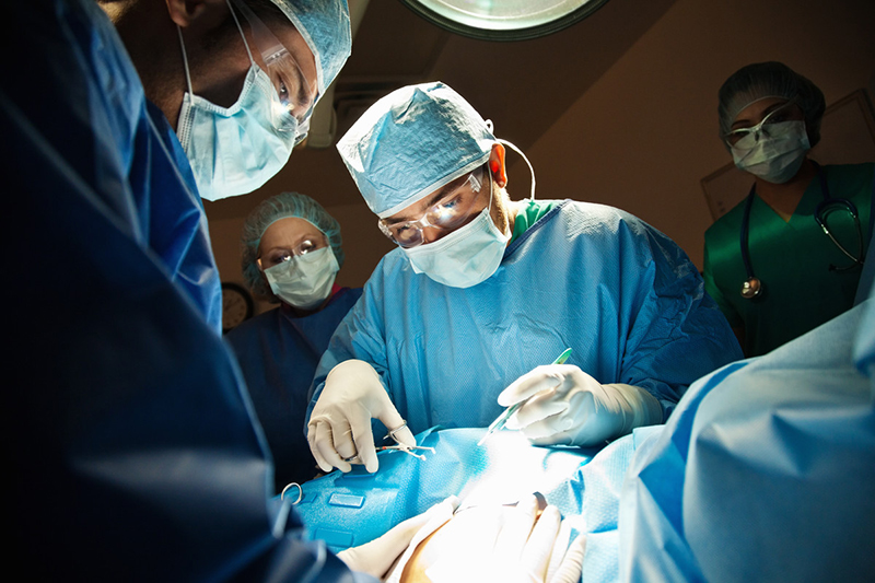 Medical professionals in surgical garb surround the draped torso of a pregnant person, while another medical professional holds surgical instruments, preparing to perform surgery.