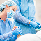 Health care worker in surgical garb holds hand of pregnant person lying on gurney. Upper torso of health care worker in surgical garb visible in background.