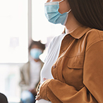 Masked medical professional writing on a desk top while a masked pregnant woman watches.
