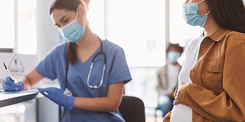 Masked medical professional writing on a desk top while a masked pregnant person looks on.