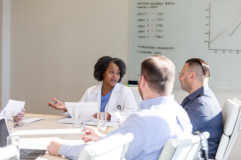 A Black woman wearing a lab coat sits at a table in a conference room speaking to two male colleagues. A whiteboard is visible in the background.