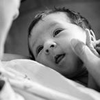 Black and white photo of a calm newborn. The baby is being held by a woman, whose face is shown out-of-focus on the left of the image. The baby is clutching the woman’s hand.