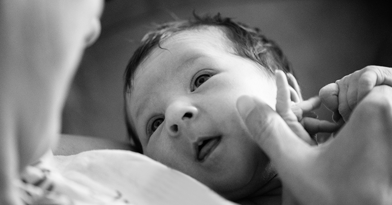 Black and white photo of a calm newborn. The baby is being held by a woman, whose face is shown out-of-focus on the left of the image. The baby is clutching the woman’s hand.