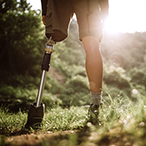 Person with a lower-leg prosthesis stands on a grassy trail. Trees and sunshine are visible in the background.