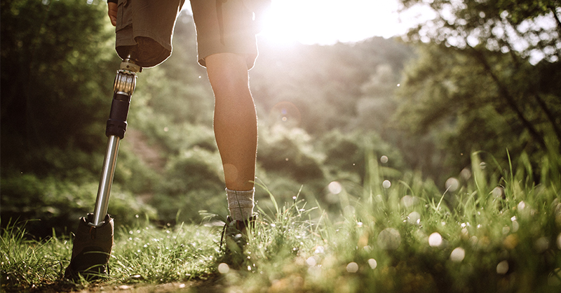 Person with a lower-leg prosthesis stands on a grassy trail. Trees and sunshine are visible in the background.